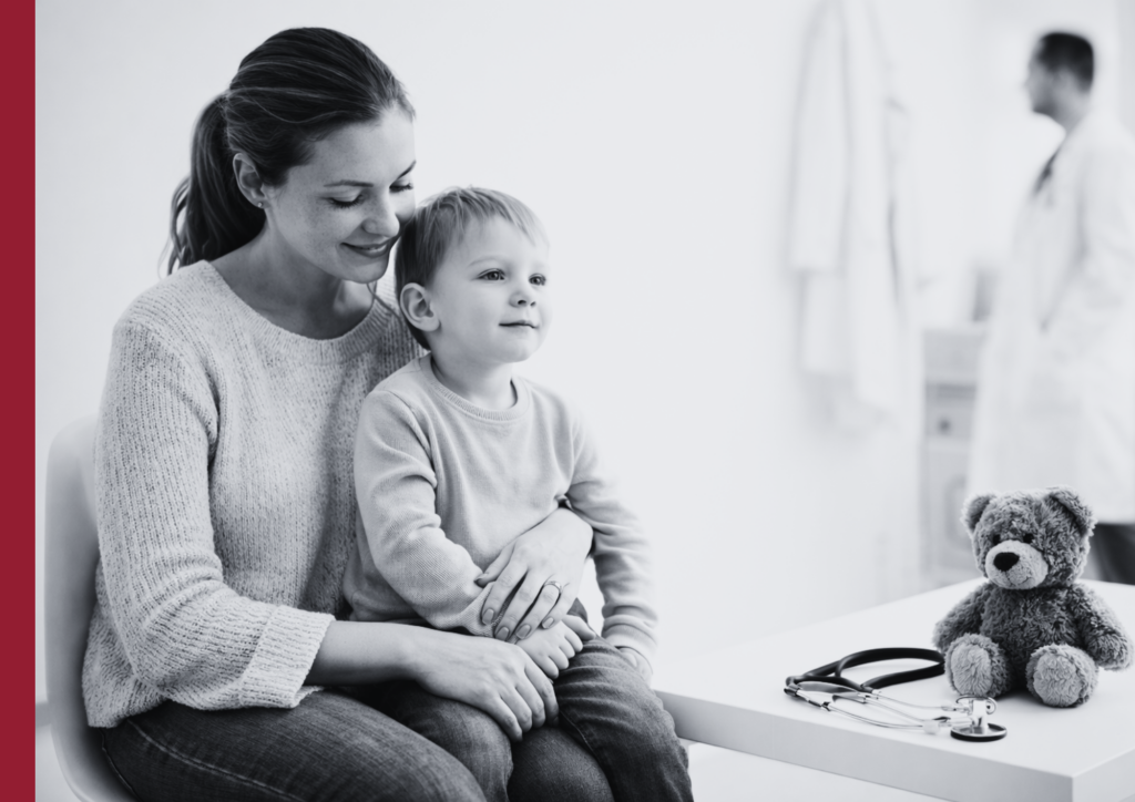 Madre con su hijo en una sala de espera pediátrica, con un peluche y un estetoscopio sobre la mesa y un médico al fondo desenfocado
